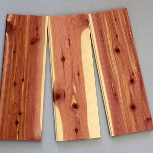 Three pieces of cedar tongue and groove siding laid out flat, showing the natural red and gold tones of the wood with visible grain patterns.